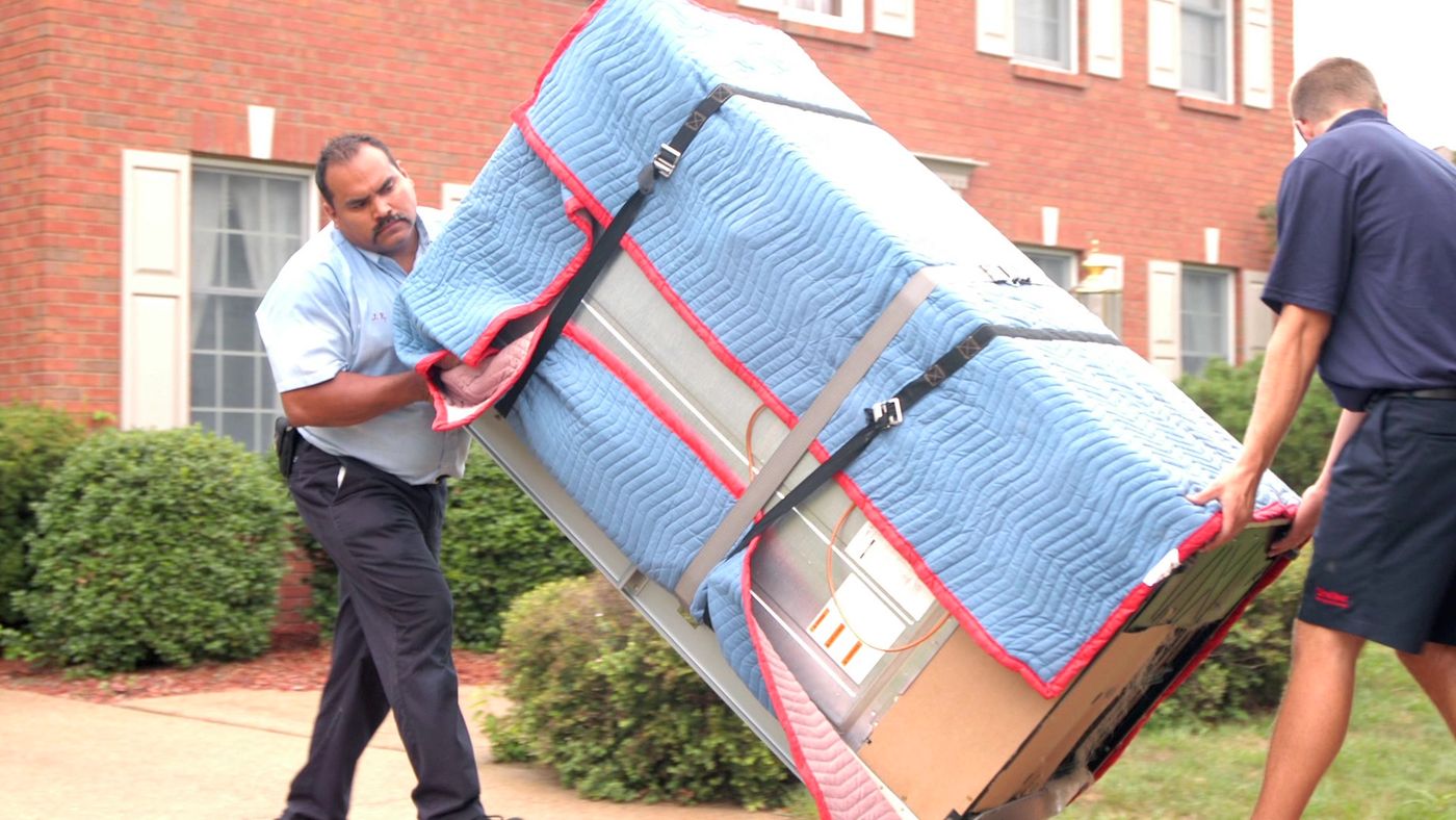 Two movers wheeling a blanket-wrapped piece of furniture on a hand truck outside a brick home