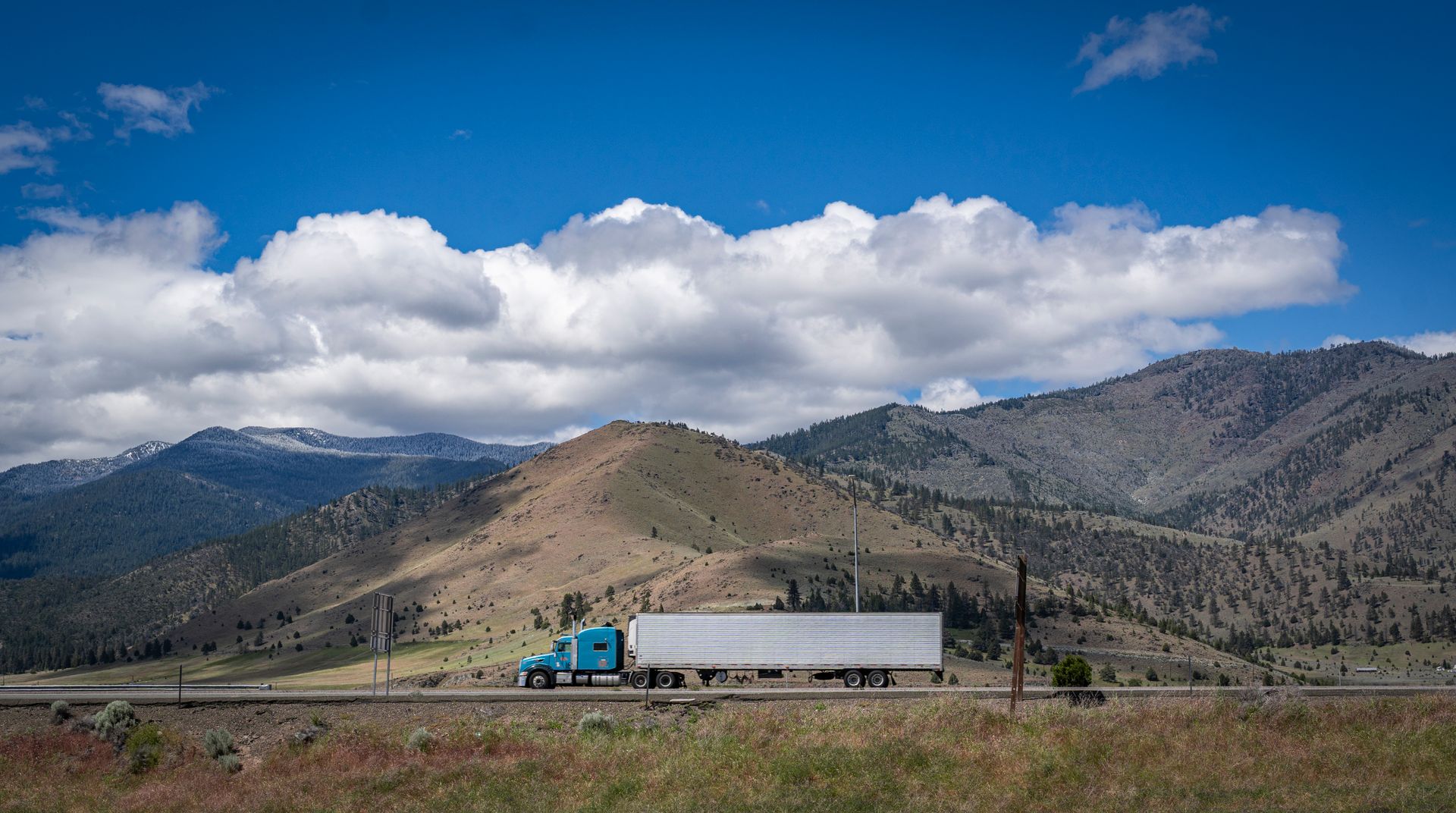 A blue semi-truck on a scenic highway with mountains and clouds