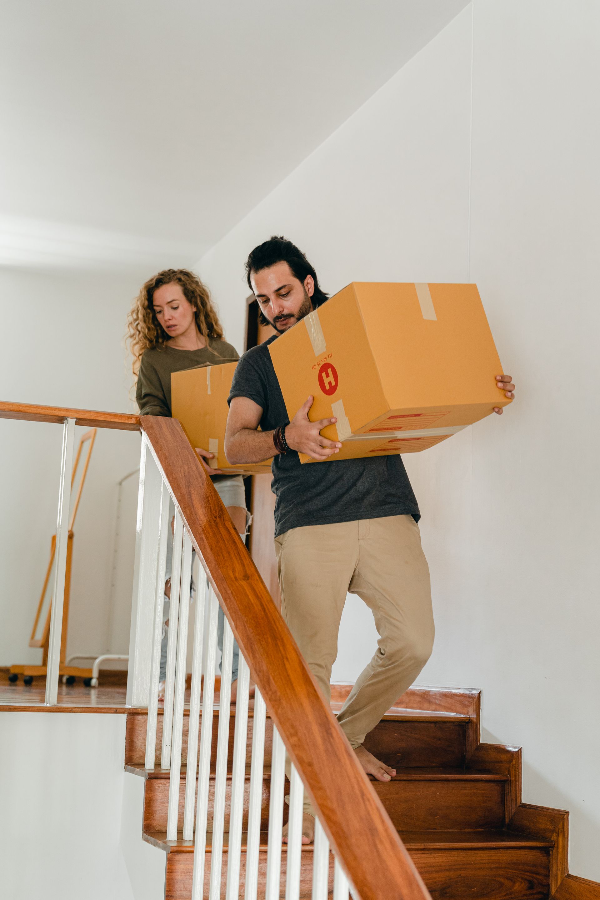 A couple carrying moving boxes down a wooden staircase in a new home